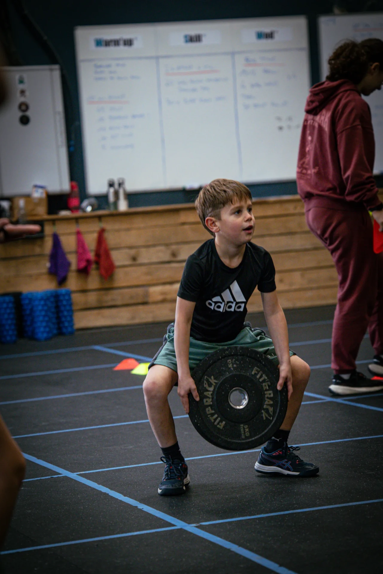 Enfant en squat avec un disque devant le tableau du WOD à la box Blue Bird